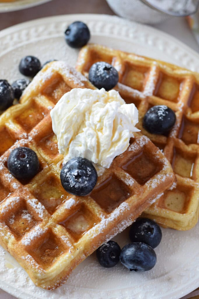Close up of waffles with whipped cream and blueberries on a white plate.