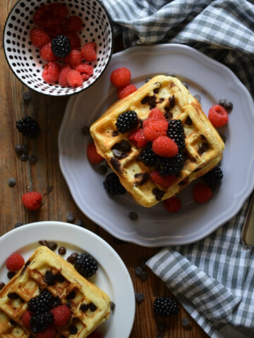 Two stacks of chocolate chip waffles on plates with berries.