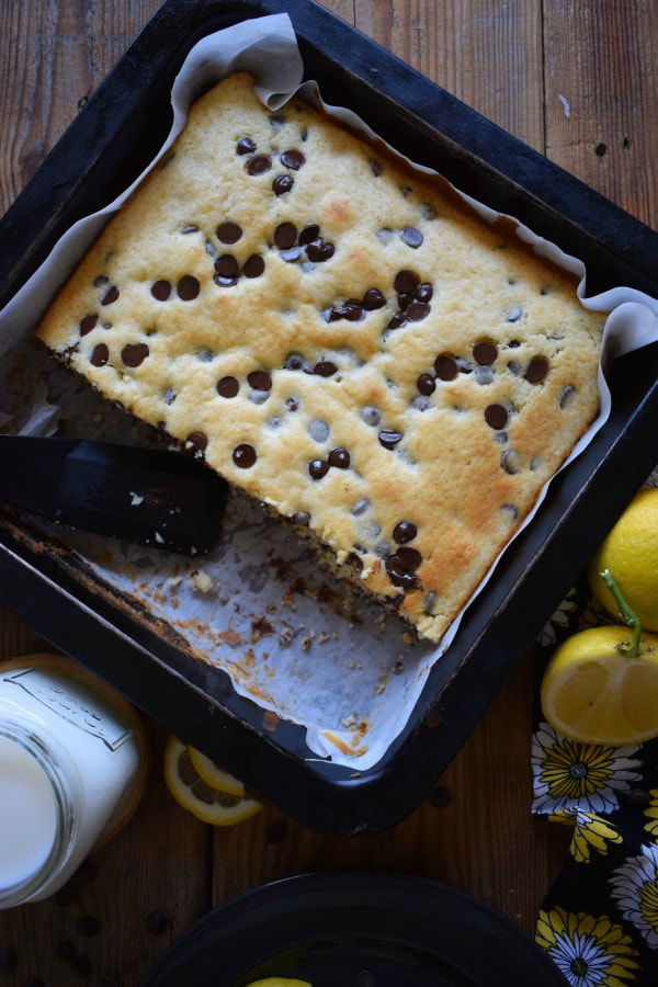 Chocolate Chip Lemon Squares in a baking tin with lemons and a glass of milk