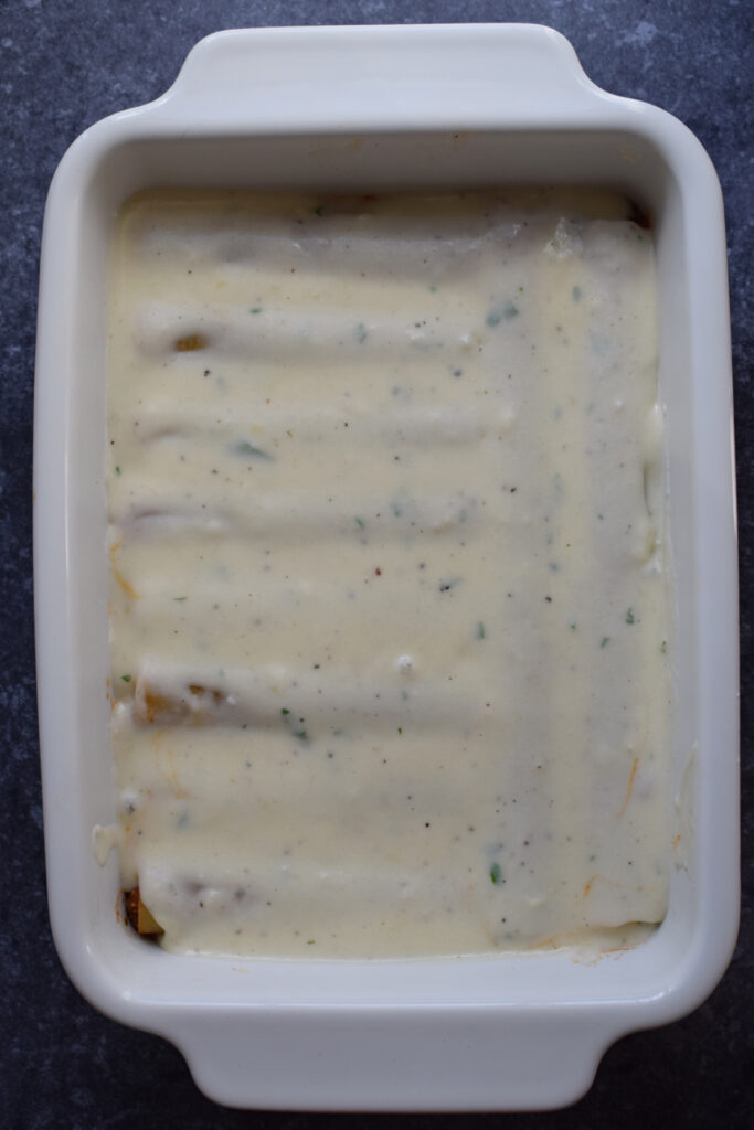 Bechamel sauce poured over cannelloni in a baking dish.