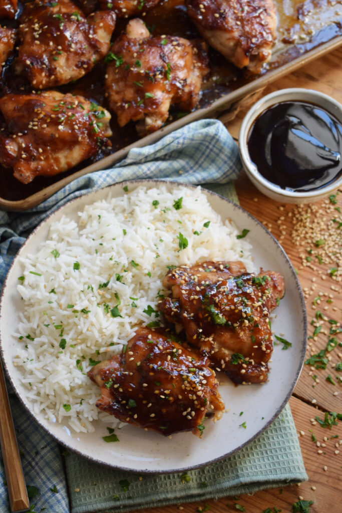 Teriyaki chicken thighs on a plate with rice and sauce on the side.