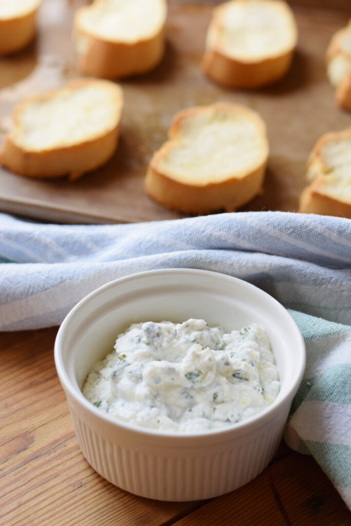 Ricotta cheese in a bowl with baguette bread slices on a baking tray.