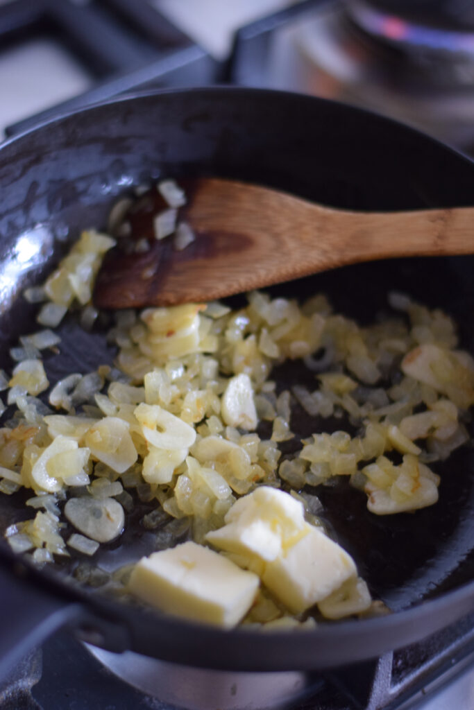 Adding butter to a skillet with onions and garlic.