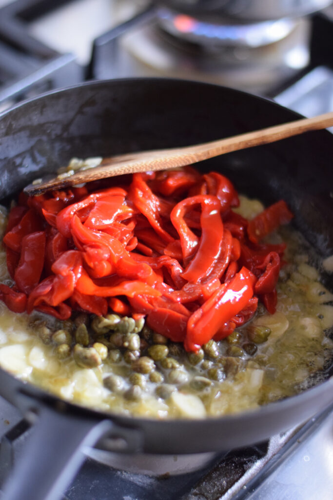 Adding roasted red peppers to a skillet with capers and onions.