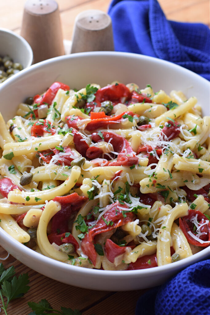 A white bowl with pasta, red peppers and capers.