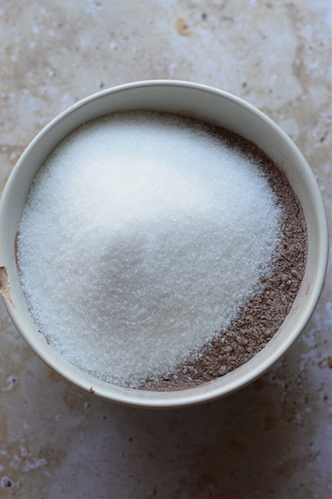 Adding sugar to dry ingredients in a bowl.