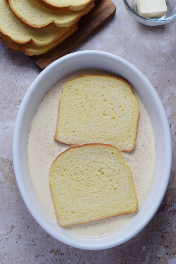 Bread slices soaking in a batter in a white bowl.