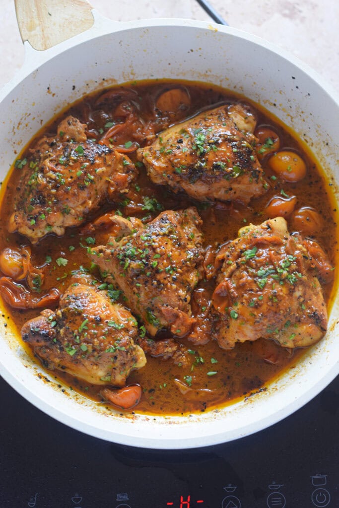 Chicken thighs cooked in a tomato broth in a white skillet.