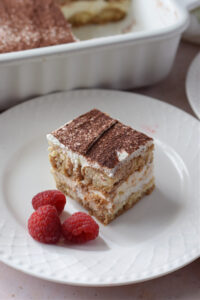 A slice of tiramisu on a white plate with raspberries.