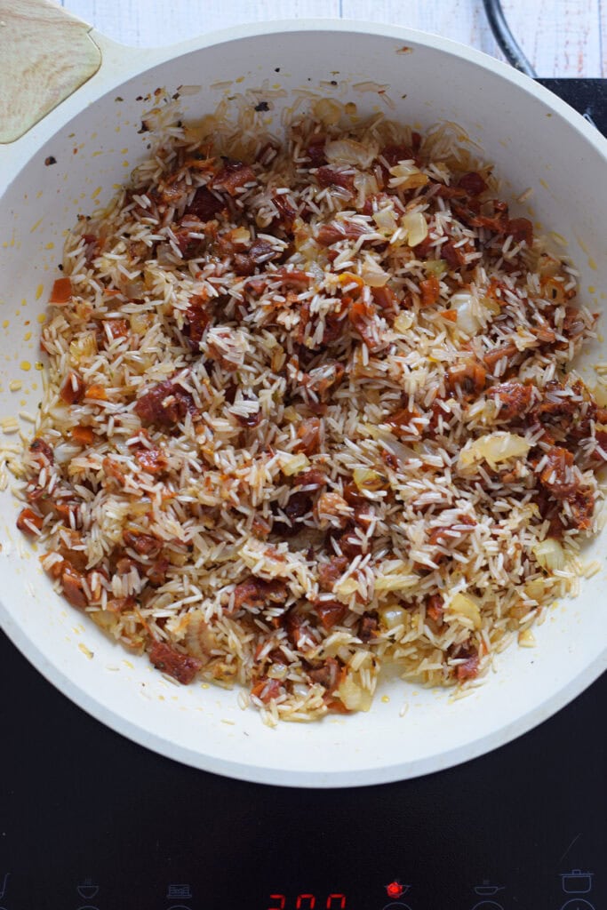 Adding rice to a skillet with onions and sun dried tomatoes.