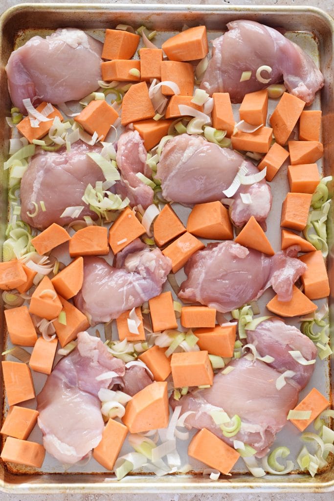 Uncooked chicken, sweet potatoes and leeks on a baking tray.
